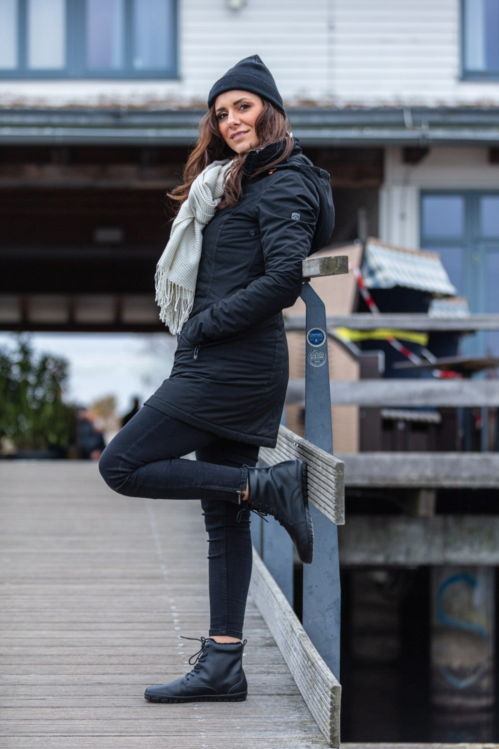 Eine Frau steht auf einem Holzsteg und lehnt an einem Geländer. Sie trägt einen schwarzen Mantel, einen Schal, eine Beanie und die QUINTIC Winter Vegan Black Schuhe von ZAQQ aus wasserabweisendem Material. Ihre schwarzen Jeans vervollständigen das Ensemble. Im Hintergrund ist ein Gebäude mit großen Fenstern und einer Holzkonstruktion zu sehen. Die Szene wirkt ruhig und lässig.