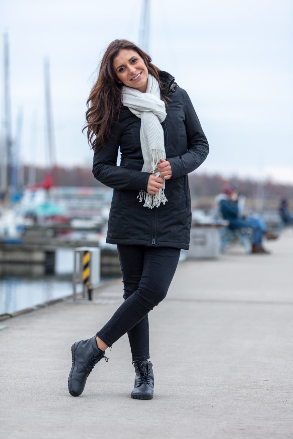 Eine Frau steht lächelnd in ihrer schwarzen Jacke und Hose auf einem Gehweg in einem Yachthafen. Ihre Füße stecken bequem in den QUINTIC Winter Vegan Black-Schuhen von ZAQQ aus wasserabweisendem Material. Ihr weißer Schal sorgt für einen Kontrast, während sie die Hände vor sich verschränkt. Ihr langes braunes Haar weht sanft, hinter ihr liegen Boote, die unter dem bewölkten Himmel angedockt sind.