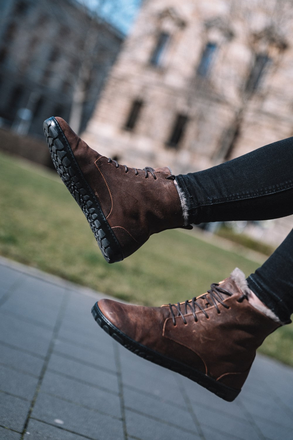 Eine Person trägt QUINTIC Winter Velours Brown Waterproof-Stiefel von ZAQQ, mit dunkelbraunen Schnürsenkeln, schwarzer Sohle und Lammfell-Futter, ideal für den Winter, und springt auf einem gepflasterten Gehweg herum. Die schwarze Hose unterstreicht den Barfußschuh-Stil. Im Hintergrund erinnern ein verschwommenes historisches Gebäude und grünes Gras an eine städtische Parkkulisse.