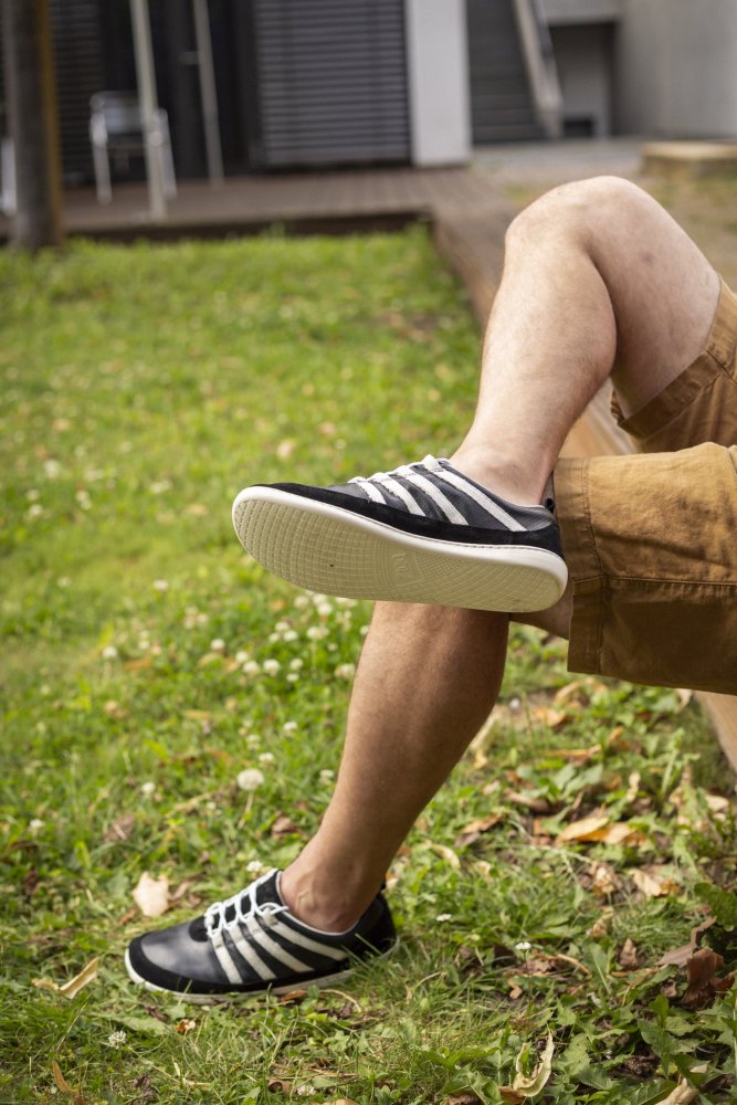 Eine Person entspannt sich mit gekreuzten Beinen auf einer Rasenfläche und trägt die SPARQ Low Black White-Sneakers von ZAQQ und Khaki-Shorts. Die friedliche Umgebung bietet eine bezaubernde Terrasse, umgeben von üppigem Grün und einem einladenden Stuhl.