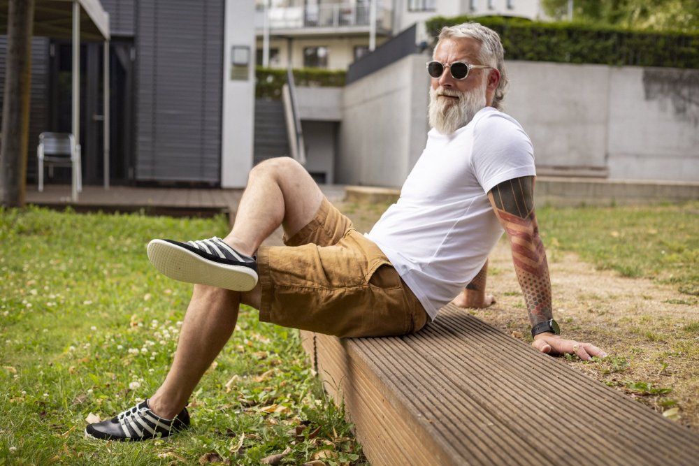 Ein bärtiger Mann mit Sonnenbrille entspannt sich draußen auf einer Holzplattform. Bekleidet mit einem weißen T-Shirt, braunen Shorts und eleganten SPARQ Low Black White Sneakers von ZAQQ mit weißen Schnürsenkeln sitzt er vor der Kulisse eines modernen Gebäudes und leuchtendem Grün.