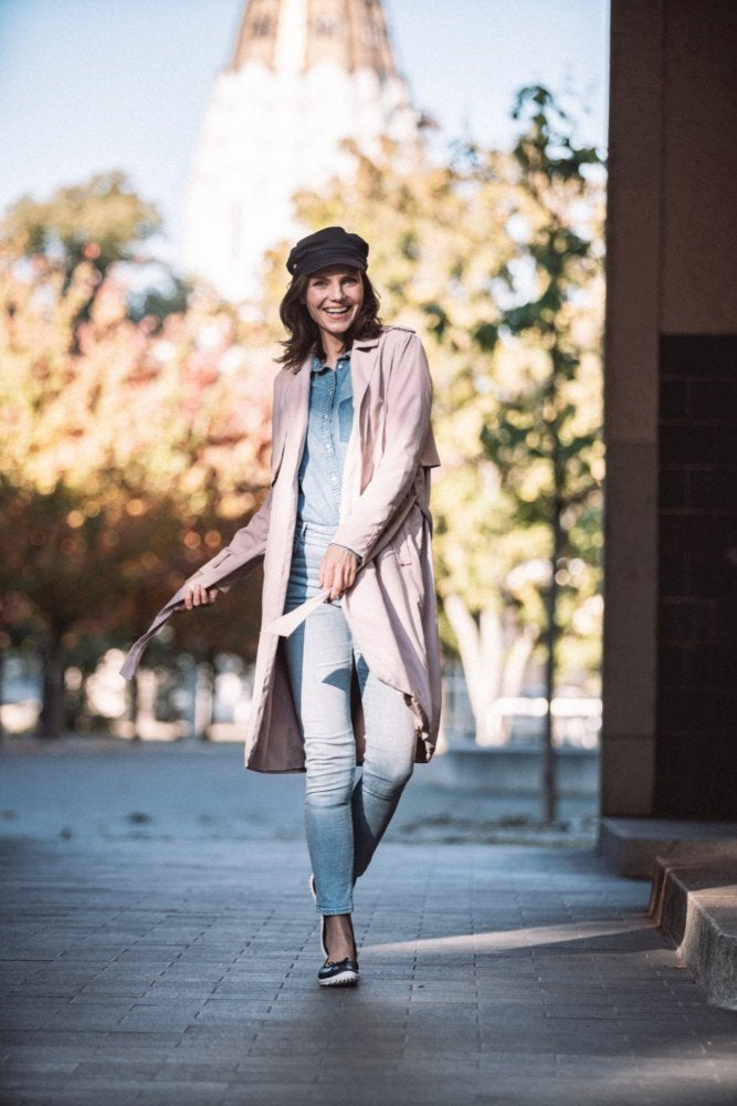 Eine Frau geht selbstbewusst durch eine Stadtstraße. Sie trägt einen langen rosa Mantel, ein hellblaues Jeans-Outfit und eine schwarze Kappe sowie schicke dunkelblaue Schuhe von ZAQQ SUQAR. Sie lächelt strahlend, im Hintergrund sieht man Herbstbäume und ein unscharfes Gebäude.