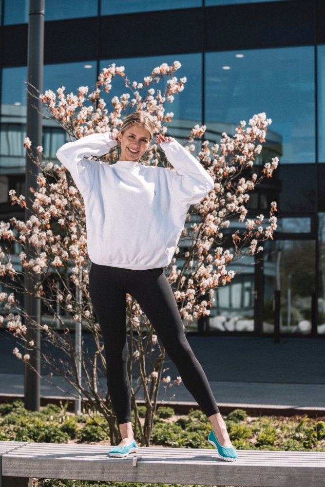 Eine Person in einem weißen Sweatshirt und schwarzen Leggings posiert draußen auf einer Holzbank und trägt ihr Lieblingspaar SUQAR Heaven von ZAQQ. Hinter ihr sind ein blühender Baum und ein modernes Glasgebäude. Sie lächeln, genießen die Atmungsaktivität und halten sich mit den Händen die Haare.