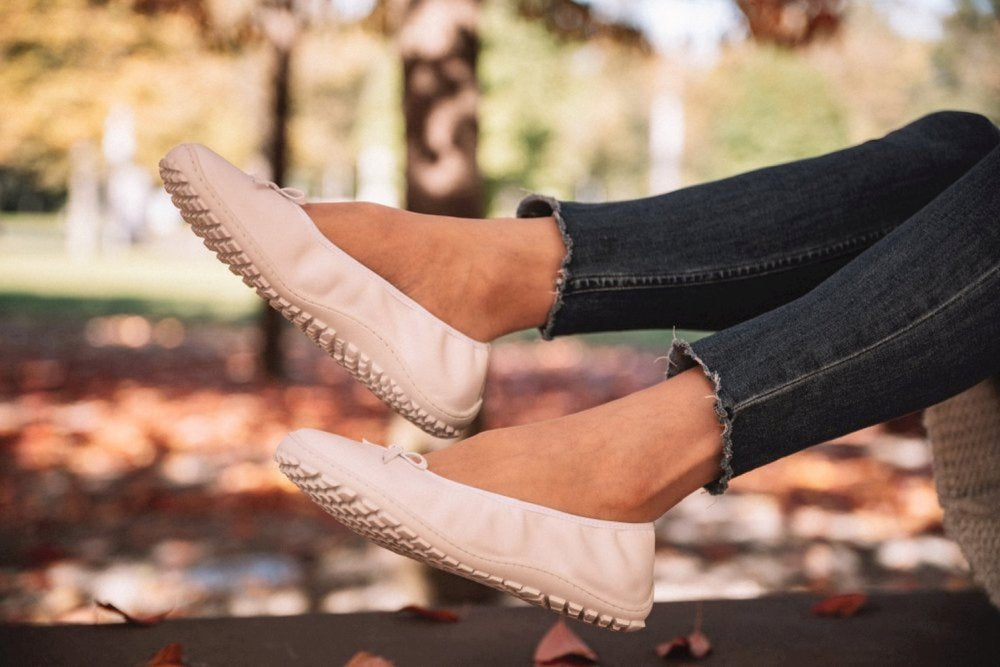 Eine Person, die die weißen SUQAR-Schuhe von ZAQQ trägt, sitzt draußen. Die Person trägt dunkle Jeans mit ausgefranstem Saum und im Hintergrund sind Bäume zu sehen, auf deren Boden Herbstblätter verstreut sind.