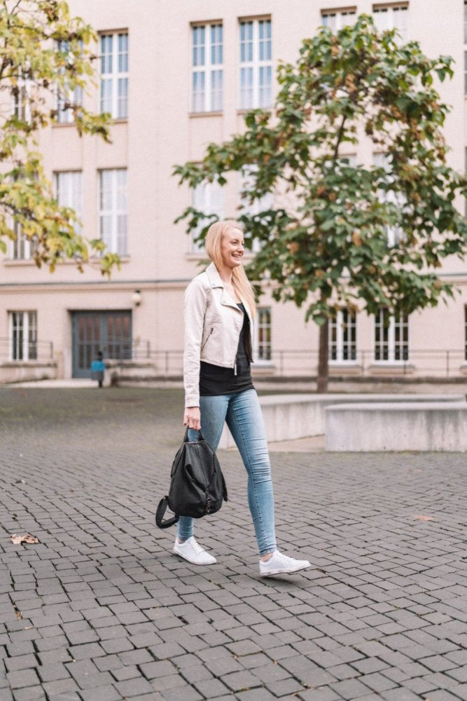 Eine Person mit langen Haaren schlendert einen Kopfsteinpflasterweg in der Nähe eines Gebäudes mit großen Fenstern entlang. Sie trägt eine leichte Jacke, Jeans und die bequemen ZAQQ TAQQ Nappa Lightblue Schuhe und trägt einen Rucksack. Bäume mit grünen Blättern dienen als Hintergrund.