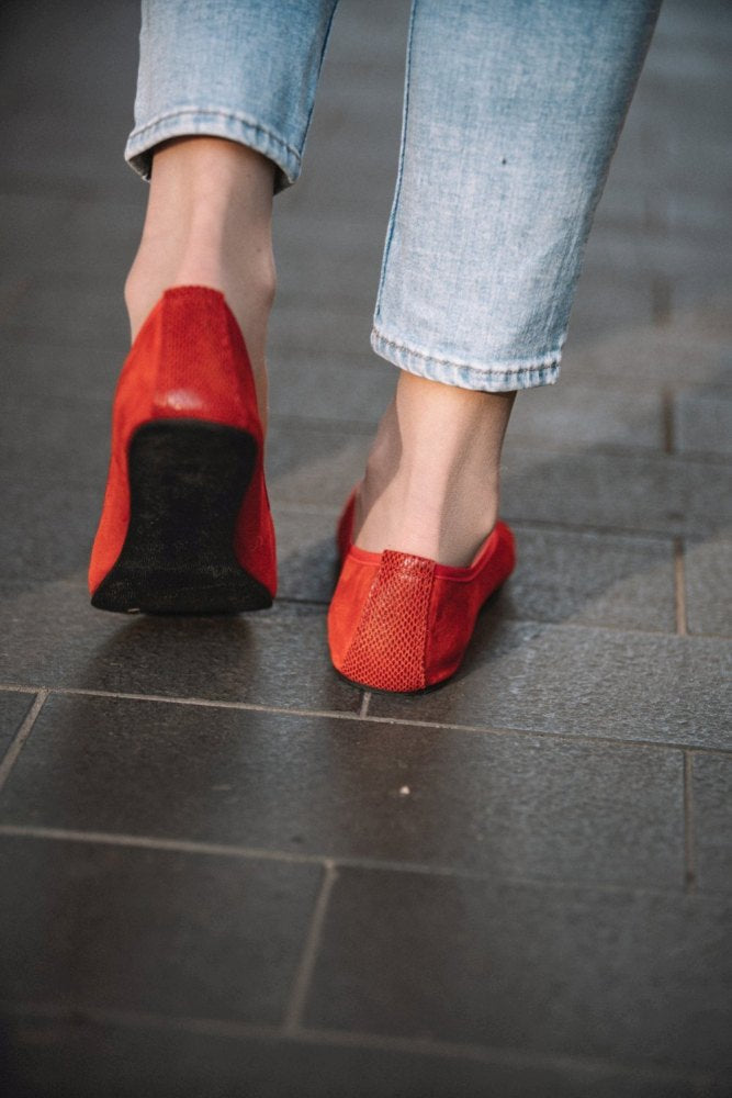 Eine Person steht auf einem grau gefliesten Boden und trägt hellblaue Jeans und ein Paar TWIST Velours Red-Schuhe von ZAQQ. Das Bild zeigt sie von hinten, mit Fokus auf ihre Füße und den unteren Teil der Jeans, wodurch die Eleganz dieser Schuhe im Ballerina-Stil mit einer glatten Kautschuksohle hervorgehoben wird.